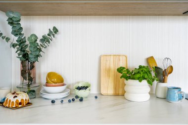 Beyaz mermer tezgah, içinde fesleğen, yabanmersini, ahşap kesme tahtası, kaseler, tabaklar ve pasta var. Yeşilci mutfak otları, çatal bıçakları ve yemek masasının üzerindeki dekorlar fotokopi odasının duvarına karşı.