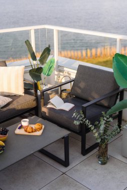 Okyanusa bakan bir avlu. Çatı terası ve açık mobilya seti. Kanepede gri yastıklı balkon ve yumuşak sandalyede kitap. Koltuk ve kanepeye karşı kahve fincanı ve hamur işi.