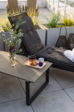 Kahvaltının yanındaki sehpaya yakın rahat bir dinlenme odası, bir kase meyve ve terasta bir kruvasan. Gri yastıklı güneşli bir güneş yatağı, havuz için kuru havlu.