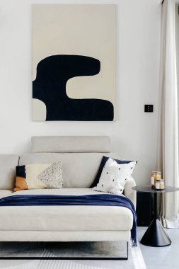 Geniş bir oturma odasında dekoratif yastıklarla süslenmiş nötr renkli bir kanepe bulunur. Kanepenin üstünde soyut bir sanat eseri asılı, şık dekoru tamamlıyor, rahat bir ambiyans sergiliyor..