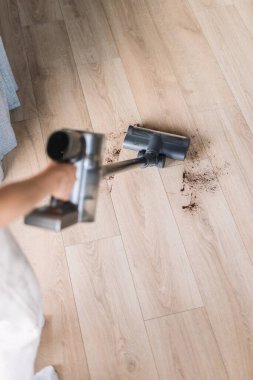 Bir insanın yüksek açılı görüntüsü elektrikli süpürge hortumunu tutar, oturma alanındaki ahşap zeminden toz ve döküntüleri kaldırır, temiz ve düzenli bir ev görüntüsü verir.