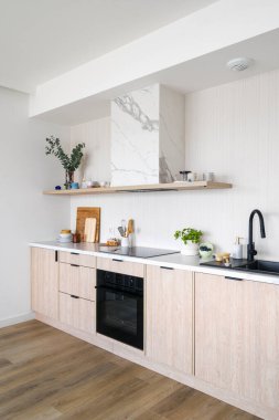 Modern mutfak cam seramik soba, elektrikli fırın, siyah lavabo ve tezgahın üstünde ahşap bir raf. Odasında beyaz mermer arka plan ve hafif ahşap dolaplar olan modern bir daire.