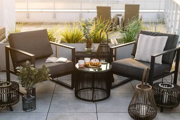 Balkonda gece fenerlerine yakın iki modern teras sandalyesi. Çatıdaki cam masanın yanındaki koltuklarda yeşil saksı bitkileri var. Arka planda şehir manzaralı konut binası