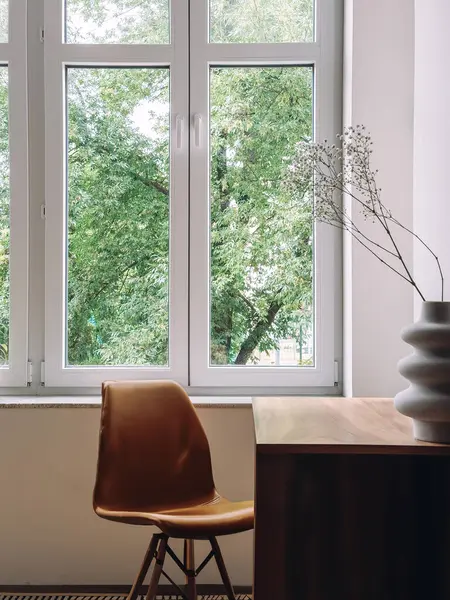Çalışma masasının dikey görüntüsü ve oturma odasındaki geniş pencerenin yanındaki ergonomik sandalye. Doğal ışık alanı masa, koltuk, ahşap mobilya ve minimalist iç tasarım ile doldurur.