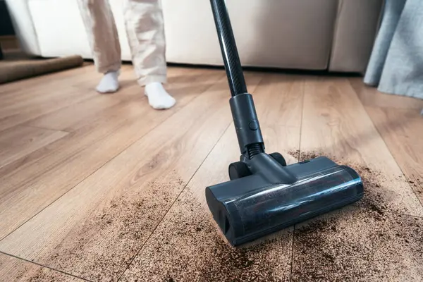 Bir insan, rahat bir oturma odasındaki ahşap zeminden toprak ve tozu temizlemek için elektrikli süpürge kullanıyor. Yakın çekim, fırçaya odaklan