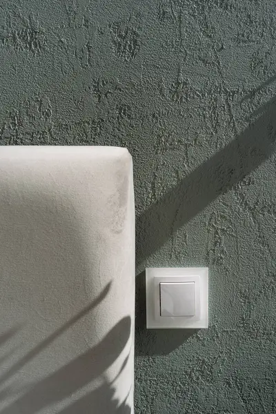 Yatağa yakın yeşil desenli bir duvara monte edilmiş beyaz ışık düğmesinin yakın çekim ve dikey görüntüsü gri yatak başlığıyla. Elektrik teçhizatı ve yumuşak yatak odası mobilyalarına odaklan.