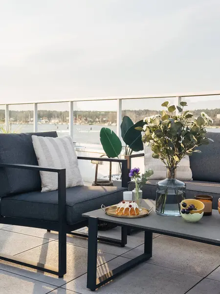 Çatı terası. Balkondaki gri koltukta dekoratif yastık rahat kanepeye ve yemekle dolu masaya yakın. Şehir manzaralı modern çatı terası, mobilyalar, saksı bitkisi ve bir tepsi şeker.