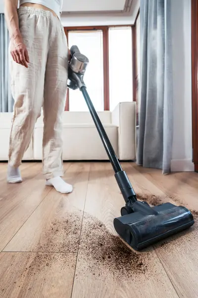 Kadın, iyi aydınlatılmış oturma odasındaki ahşap zemini temizlemek için elektrikli süpürge kullanıyor. Geniş pencereleri ve rahat bir kanepesi var.