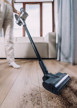 Parlak ve havadar bir oturma odasındaki ahşap zemini temizlemek için kablosuz elektrik süpürgesi ve arka planda rahat bir kanepe kullanan biri.
