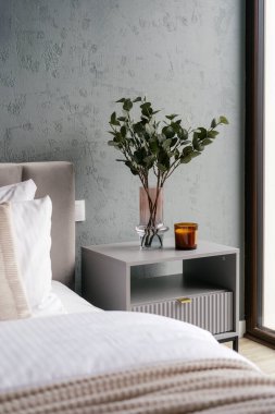 Otel odasındaki yatağın yanında mum ve ev dekorasyonuyla komodinin yan görüntüsü. Yatak odasının içinde komodinin üzerinde okaliptüs bitkileri var. Sessiz yaşam, stil mekanları kavramları