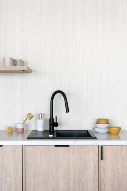 Modern bir mutfakta siyah musluk beyaz fotokopi duvarına karşı duruyor. Tezgah temiz ve minimalist. Lavabonun üstünde küçük bir raf var.