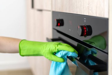 Eldivenli bir elin, siyah fırın kapısını mavi bir bezle silerken yakın plan çekimi. Kadın, mutfak aletlerini toz ve pislikten temizlemek için yumuşak bez kullanıyor.