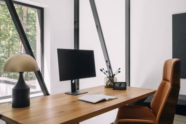 Çağdaş çalışma alanı parlak ahşap bir masa, bilgisayar monitörü ve zarif bir dekor içerir. Masa lambası ve büyük pencerelerle çevrili bir bitki olan oda ev ofisinin manzarasını sunuyor.