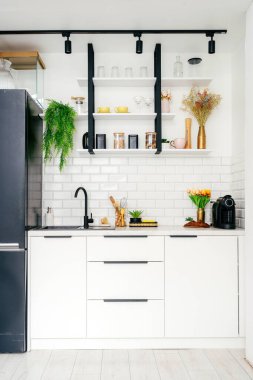 Modern bir mutfak, gösterişli beyaz dolaplar, siyah donanım, mutfak gereçleri ve dekoratif bitkiler içerir ve evde yemek pişirmek için davet edici bir alan yaratır. İç mekanın dikey görüntüsü