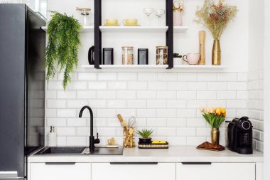 Beyaz modern mutfak tezgah ve raflarda bitkiler, mutfak gereçleri ve dekoratif kavanozlarla süslü bir iç tasarım sergiliyor. Taze çiçekler, davetkar ve düzenli bir atmosfer vurgulanıyor.