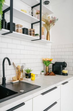 Gösterişli beyaz bir mutfakta siyah armatürler, duvar fayansları ve dekoratif aksanlar, tezgahta taze çiçekler ve şık mutfak eşyaları sergileniyor. Dairenin iç kısmının dikey görüntüsü