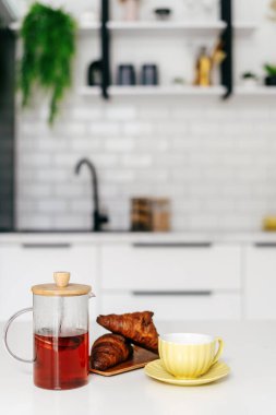 Şık bir mutfakta çay, sarı bardak ve ahşap tepside tereyağlı kruvasanlarla dolu cam bir çaydanlık bulunur. Lezzetli yemek kavramları ve evdeki ılık sabah atmosferi.