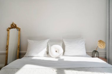 Sakin bir yatak odasında, düzgün yapılmış bir yatakta beyaz yastıklar, klasik bir ayna ve doğal ışıkla hafifçe aydınlatılmış kurumuş çiçekli şık bir vazo bulunur. Otel odası.