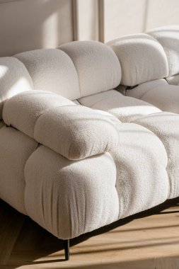 Rahat bir oturma odasında, modern tasarımı ve dikey çekimi olan yumuşak, beyaz, peluş bir kanepe sergileniyor. Ilık güneş ışığı filtreleri, şık mobilyaları vurgulamak ve uzayın davetkar atmosferi.