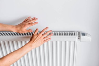 Eller, soğuk oturma odasında sıcaklık sağlayan beyaz bir elektrikli ısıtıcıya uzanıyor. Bu ısıtma çözeltisi evdeki konfor ve enerji verimliliğini artırır..