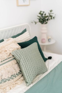 Sakin bir yatak odası dekoratif yastıkları çeşitli doku ve renklerde, yumuşak bir yatak örtüsü ve yeşil vazo ile tamamlanmış, barışçıl bir ortam yaratıyor..