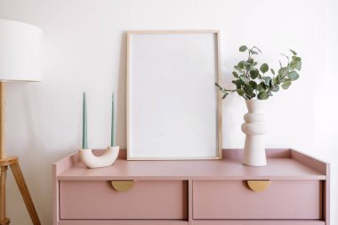 Şık bir oturma odasında boş bir çerçeve, dekoratif vazolar, mumlar ve yemyeşil çiçeklerle süslenmiş minimalist bir konsol masası sergileniyor. Konfor ve çağdaş ev estetiğini vurguluyor..