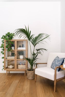 Modern oturma odasının dikey çekimlerinde rahat bir beyaz koltuk, dekoratif eşyalar sergilenen ahşap bir dolap ve davetkar alanı geliştiren yemyeşil bitkiler yer alıyor..