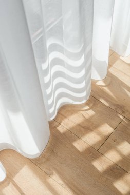 Pencere tülü. Güneş ışığı beyaz perdelerin arasından akar, zarif bir dairenin sıcak ahşap zeminine yumuşak gölgeler bırakır, odada sakin ve davetkar bir atmosfer yaratır.