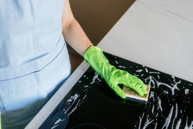 Yeşil eldivenli bir kadın şık bir mutfakta indüksiyon sobasını temizliyor. Tezgah sabun köpüğüyle kaplı. Bu da düzenli bir ev ortamının korunmasına olan bağlılığını gösteriyor..