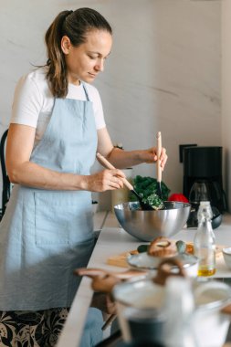 Modern bir mutfakta, bir kadın salata kasesinde taze sebzeleri karıştırmaya odaklanır, evde yemek pişirirken sağlıklı bir yaşam tarzını teşvik eder. Tezgah çeşitli malzemelerle güzelce düzenlenmiş..
