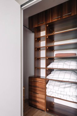 Gardırop yatak odasında. Zarif dolabın yan tarafında ahşap raflar, düzgünce katlanmış çarşaf ve havlular, odayı modern estetik olarak geliştirecek minimalist bir tasarım sergileniyor..