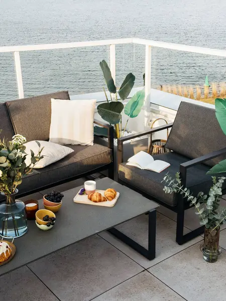 Gri dış mobilya ve su manzaralı modern verandanın dikey görüntüsü. Konforlu kanepe, sehpa ve sandalyeli balkon. Tatil için çatı terası olan bir apartman. Otel tesisi
