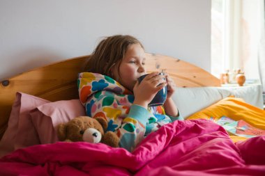 Kid lying in bed with pink blanket. Little girl with cold in scarf and blanket with hot beverage in hands near teddy bear at home