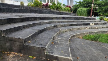 Doğal taştan yapılmış merdivenler, yeşil yapraklı Ketapang ağaçlarıyla dolu bir bahçede estetik olarak düzenlenir ve parlak bir atmosferde bulutlu mavi gökyüzü arkaplanı vardır..