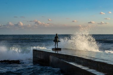 Paphos, Kıbrıs - 23 Aralık 2023: Paphos gezinti güvertesine büyük deniz dalgaları düştü. İskelede küçük bir balıkçı heykeli.