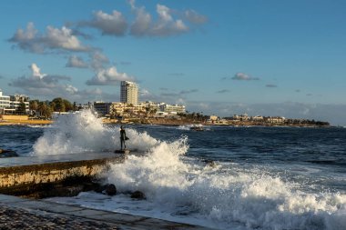 Paphos, Kıbrıs - 23 Aralık 2023: Paphos gezinti güvertesine büyük deniz dalgaları düştü. İskelede küçük bir balıkçı heykeli.