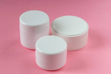 Pembe arka planda üç beyaz kozmetik kavanozu duruyor. Kozmetoloji, tıp, güzellik, sağlık ve bakım