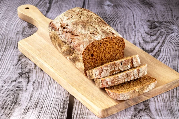 On a cutting board, a loaf of dark bread with pieces on a wooden background