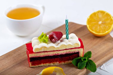 A piece of raspberry cake with fluffy cream and a candle on a wooden board. Nearby are mint leaves, lemon peel, orange and a cup of tea