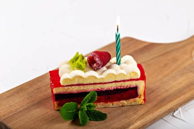 A piece of raspberry cake with fluffy cream with a burning candle and mint leaves on a wooden board