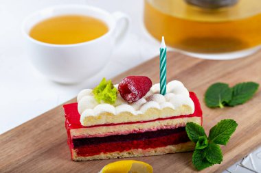 Raspberry cake with raspberry berry on fluffy cream and candle on wooden board. Mint leaves, lemon peel and tea in cup and teapot nearby
