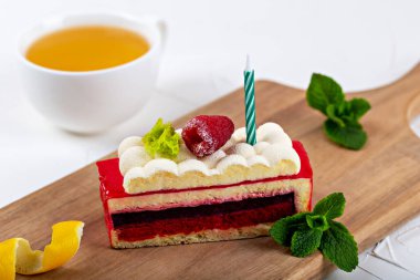 Fresh appetizing raspberry cake with berry and candle on fluffy cream on wooden board. Mint leaves, lemon peel and cup of tea are nearby