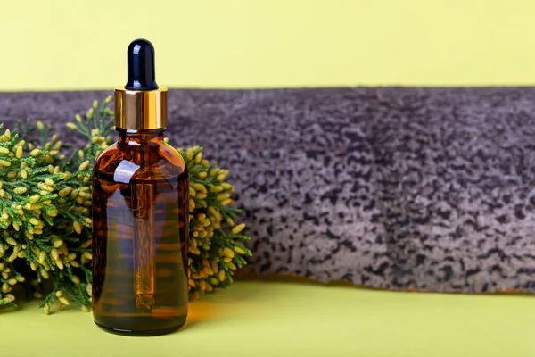 Glass cosmetic amber bottle, juniper branch and tree bark on yellow background close up. Organic cosmetics. Natural care