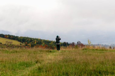 Bulutlu dağlarda telefonuyla fotoğraf çeken yabancı bir adam. Güzel sonbahar manzarası, bulutlarda gökyüzü, çimenler ve ağaçlar. Turizm ve seyahat
