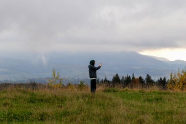 Ceketli ve pantolonlu adam dağların arasında duruyor ve telefonuyla fotoğraf çekiyor. Güzel sonbahar manzarası, bulutlarda gökyüzü, çimenler ve ağaçlar. Turizm ve seyahat