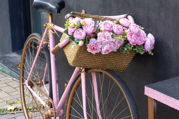 Pink bicycle with basket of pink peony flowers stands near black wall. Love, romance and relationships