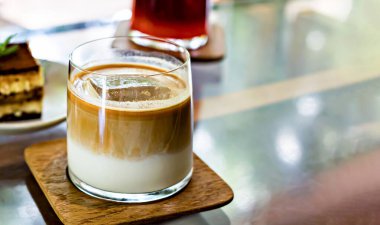 Süt katmanlı buzlu kahve ve ahşap bardak altlığında espresso. Arkaplanda da tatlı. Sofistike kahve molası için ideal.