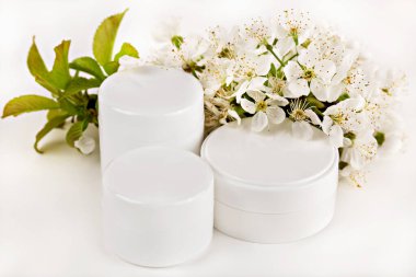 Modelleme. Kozmetik krema kavanozları beyaz çiçekler ve taze yeşil yapraklarla düzenlenir. Krem doğal güzellik cildini ön plana çıkarır. Zarif organik cilt damgalama kullanımı için kavramsal kozmetik kremi