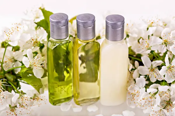 Üç şeffaf cilt şişesi narin beyaz çiçekler arasında düzenlenir. Taze çiçekli arka plan doğal, organik güzellik ve refah kavramını geliştirir, saflığı ve kendine bakımı sembolize eder.