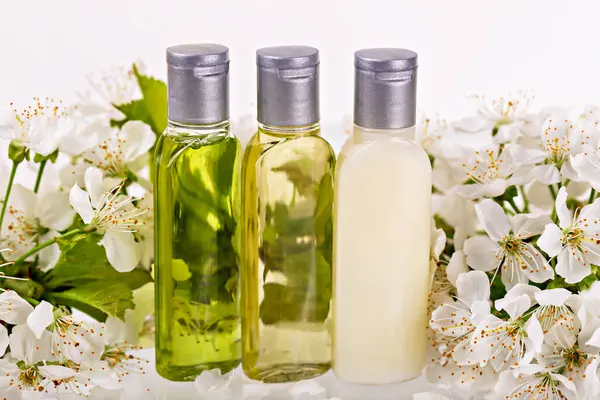 Gümüş kapaklı şeffaf üç şişe beyaz çiçeklere karşı hazırlanmış doğal cilt yağları içerir. Taze çiçekli arka plan organik güzelliği, saflığı ve bütünsel kendine bakımı vurgular.
